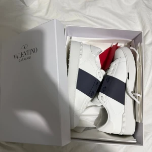 Valentino Open - Snygga Valentino Garavani Open sneakers i vitt skinn med bred mörkblå rand över sidan. Klassisk rund tå, platt sula med mönster och subtila nitar på hälen. Guldlogga på plösen och innersulan. Perfekt för dig som gillar exklusiv streetstyle.