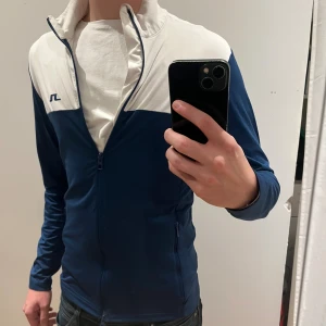 Blåvit vindjacka från J.Lindeberg - Snygg blå och vit vindjacka från J.Lindeberg med dragkedja framtill och ståkrage. Jackan har lång ärm och en cool kontrastdesign med logga på bröstet och ett vertikalt J.Lindeberg-tryck på ryggen. Skriv vid frågor ! Skick 9/10