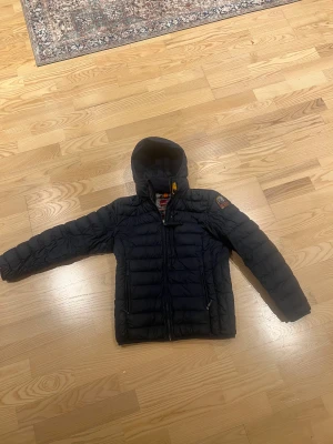 Mörkblå dunjacka Parajumpers  - Snygg mörkblå dunjacka från Parajumpers med huva och coolt patch på ärmen. Jackan har quiltad design, dragkedja framtill och två fickor med zip. Perfekt för kalla dagar och har en lätt, sportig känsla. Insidan är grå och jackan har orange detaljer. Passar folk från 158-164