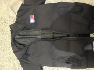 Svart pufferjacka Tommy Hilfiger - Svart pufferjacka från Tommy Hilfiger med broderad logga på bröstet och stor text på ryggen. Jackan har dragkedja framtill, ribbade fickor och klassisk krage. Perfekt för kalla dagar och har en clean, urban vibe.