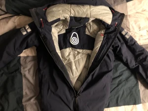 Mörkblå Sail Racing dunjacka - Säljer en mörkblå dunjacka från Sail Racing med grått foder och coola logodetaljer på ärmar och huva. Jackan har huva, dragkedja framtill och flera praktiska fickor. Perfekt för kalla dagar och har en sportig look med snygga reflexdetaljer.