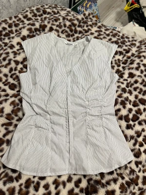 Vit randig blus från Weekday - Säljer en vit blus med svarta tunna ränder från Weekday i storlek 36. Blusen har v-ringning, ärmlös design och snygga rynkade detaljer i midjan som ger en cool siluett. Stängs med hakar framtill för en unik touch. Perfekt för dig som gillar stilrena och trendiga plagg.