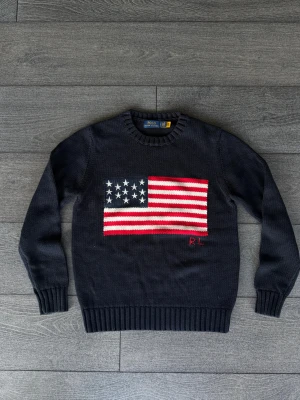 Mörkblå stickad tröja Polo Ralph Lauren - Mörkblå stickad tröja från Polo Ralph Lauren med amerikansk flagga broderad på bröstet och RL-detalj i rött. Klassisk rund halsringning och ribbade muddar. Perfekt för dig som gillar preppy stil och vill sticka ut med en ikonisk look.