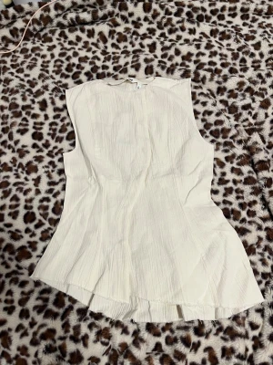 Vit blus med knappar från Weekday - Säljer en snygg vit blus från Weekday i storlek 36. Blusen är ärmlös och har en lätt strukturerad yta med knappar framtill. Den har en figurnära passform och lätt utsvängd nederkant, perfekt för dig som gillar stilrena och moderna plagg.