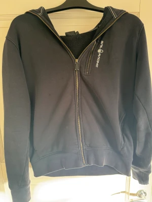 Svart hoodie från Sail Racing, M - Svart hoodie från Sail Racing i storlek M. Tröjan har hel dragkedja framtill, broderad logga på bröstet och en stor huva. Tillverkad i mjuk bomullsmix med ribbade muddar och en extra dragkedja vid bröstet. Perfekt för chill dagar.