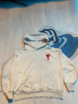Ami Paris hoodie - Säljer en beige hoodie från Ami med broderat rött hjärta och A på bröstet. Tröjan har huva, känguruficka och ribbade muddar. På ryggen finns ett diskret broderat AMI-logo. Perfekt för dig som gillar stilrena och snygga streetwear-plagg. Pris ej hugget i sten