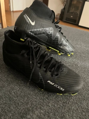 Nike Air Zoom svarta fotbollsskor - Säljer ett par svarta Nike Air Zoom fotbollsskor med snörning och högt skaft. Skorna har en modern design med diskreta mönster och detaljer i grått samt gula dobbar undertill. Tillverkade i syntetmaterial för bästa grepp och känsla på planen.