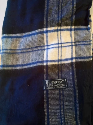 Rutig blå halsduk från Burberrys - Snygg halsduk från Burberrys i 100% lammull, tillverkad i England. Den har ett klassiskt rutigt mönster i mörkblått, vitt, grått och inslag av grönt och gult. Fransen i ändarna ger en chill vibe och den är perfekt för kalla dagar.