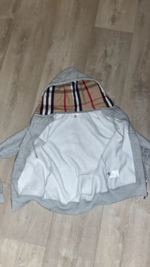 Grå hoodie med Burberry-inspirerad huva - Säljer en grå hoodie från burberry med dragkedja och snörning. Insidan av huvan har ett klassiskt Burberry-inspirerat rutigt mönster i beige, rött, blått och svart. Tröjan har långa ärmar, ribbade muddar och mjukt material som känns skönt mot huden.