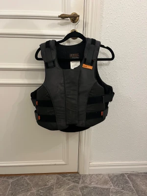 Svart säkerhetsväst Airowear L3 - Säljer en svart/grå säkerhetsväst från Airowear, modell Ladies Outlyne. Västen har justerbara kardborreband i sidorna och orange detaljer. Certifierad enligt BETA 2009 Level 3 för ridning. Materialet är slitstark syntet med ventilerande mesh på insidan. 