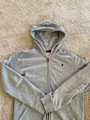 Grå hoodie från Morris Company utan snören  XS - Snygg grå hoodie från Morris Company i storlek XS. Tröjan har huva med snörning, dragkedja framtill med läderdetalj och ett mörkblått broderat emblem på bröstet. Tillverkad i mjuk bomull och har långa ärmar samt ribbade muddar vid ärmslut och nederkant.