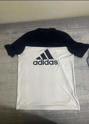 Adidas vit och svart t-shirt - Snygg t-shirt från Adidas i vit och svart med stort Adidas-tryck på bröstet. Modellen har korta ärmar och rund hals. Materialet är mjuk polyester som andas, perfekt för sport eller chill. Stilren tvåfärgad design som ger en sportig vibe.