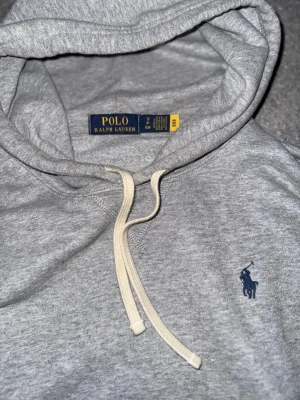 Grå hoodie från Polo Ralph Lauren - Snygg grå hoodie från Polo Ralph Lauren med klassisk blå logga broderad på bröstet. Tröjan har huva med vita snören, känguruficka och långa ärmar. Mjuk bomullskvalitet som är perfekt för chill dagar. Passformen är normal och stilen är enkel men stilren.