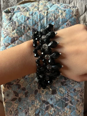 Svart pärlarmband med droppformade detaljer - Unikt svart armband med glittriga, facetterade pärlor och droppformade detaljer i svart och transparent. Armbandet har flera rader och ett smidigt metallspänne. Perfekt för dig som gillar statement-smycken och vill sticka ut med coola accessoarer.