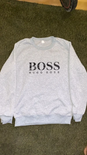 Grå sweatshirt från Hugo Boss - Snygg ljusgrå sweatshirt från Hugo Boss med svart logga framtill. Tröjan har rund halsringning, långa ärmar och ribbade muddar vid ärmslut och nederkant. Perfekt för en clean och stilren look.