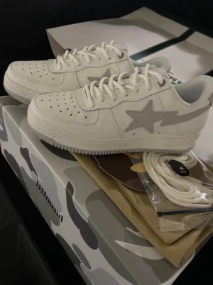 JJJJound x Bape Sta vita sneakers - Säljer ett par JJJJound x Bape Sta sneakers i vitt med grå stjärndetalj på sidan. Skorna har klassisk låg siluett, perforerad tå och platt sula. Tillverkade i skinn med vita snören och kommer i en camo-mönstrad box. Perfekt för dig som vill vara clean och stilren