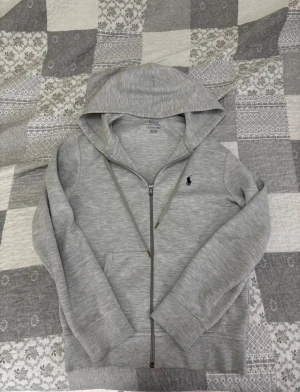 Grå hoodie från Polo Ralph Lauren XS - Snygg grå hoodie från Polo Ralph Lauren i storlek XS. Tröjan har huva med snörning, dragkedja framtill och klassisk Polo-logga broderad i svart på bröstet. Tillverkad i mjuk bomullsmix, perfekt för chill dagar. Fickor på magen och ribbade muddar vid ärmslut.
