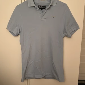 Ljusblå pikétröja från Barbour - Snygg ljusblå pikétröja från Barbour i storlek S. Klassisk krage, korta ärmar och två knappar vid halsen. Diskret Barbour-logga broderad på bröstet. Tillverkad i mjuk bomull som känns skön mot huden. Perfekt för dig som gillar stilrena och enkla plagg.