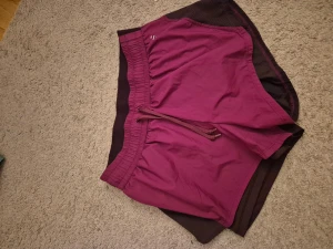 Vinröda träningsshorts med snörning - Säljer ett par vinröda träningsshorts med elastisk midja och snörning framtill. Shortsen har en loose passform och är gjorda i ett lätt syntetmaterial som andas. Perfekta för gymmet eller löprundan.