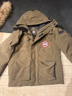 Canada Goose parkas XL med päls - Säljer en olivgrön Canada Goose parkas i storlek XL med avtagbar päls på huvan. Jackan har flera stora fickor, klassisk logga på bröstet och dragkedja framtill. Perfekt för kalla vinterdagar och har en riktigt snygg streetstil.