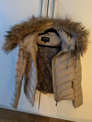 Beige pufferjacka med äkta päls  från Hollies - Snygg beige pufferjacka från Hollies med stor huva och fluffig äkta päls. Jackan har dragkedja framtill, två sidofickor med dragkedja och quiltad design. Insidan har ett coolt spetsmönster. Perfekt för kalla dagar och riktigt trendig look.