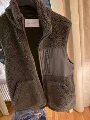 Mörkgrön fleeceväst från A Day's March - Snygg mörkgrå fleeceväst från A Day's March med hög krage och praktiska fickor. Västen har en dragkedja framtill och en extra ficka med zip på bröstet. Perfekt att slänga över en hoodie för extra värme. Mjuk och fluffig känsla.