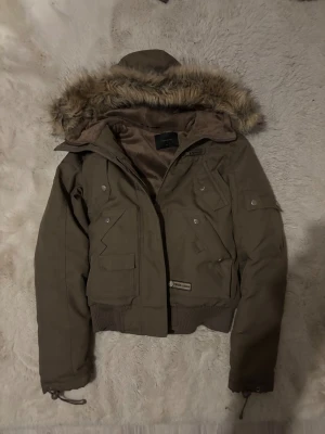 Brun pufferjacka med päls från Crystal Lodge Vero Moda - Säljer en brun pufferjacka från Crystal Lodge med fuskpäls på huvan, flera fickor med knappar och ribbade muddar. Jackan har snörning vid ärmslut och en kort, loose passform. Perfekt för kalla dagar med sin tjocka polyester och snygga detaljer.