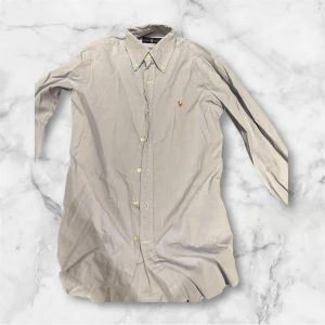 Ljus blå skjorta från Ralph Lauren - Snygg ljusblå skjorta från Ralph Lauren med klassisk krage och guldfärgade knappar. Skjortan har lång ärm och den ikoniska broderade loggan på bröstet. Perfekt för dig som gillar stilrena och tidlösa plagg. storleken är runt M.