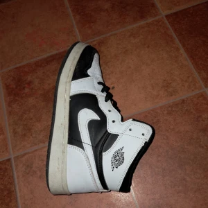 Nike Air Jordan 1 svart/vit high - Nike Air Jordan 1 sneakers i svart och vitt med klassisk high-top siluett. Skorna har snörning, perforerad tå och ikoniska Jordan-loggan på sidan. Tillverkade i skinn med platt sula och rund tå. Snygg kontrast mellan svart och vit, passar dig som gillar streetstyle.