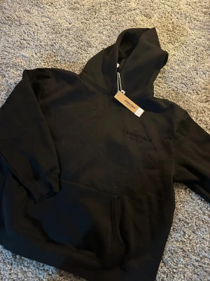 Essentials hoodie svart - Svart hoodie från Essentials Fear of God i storlek L. Tröjan har stor huva, ribbade muddar och en klassisk känguruficka framtill. Loggan 'ESSENTIALS FEAR OF GOD' är tryckt både på bröstet och ryggen. Perfekt för dig som gillar streetwear och clean design.