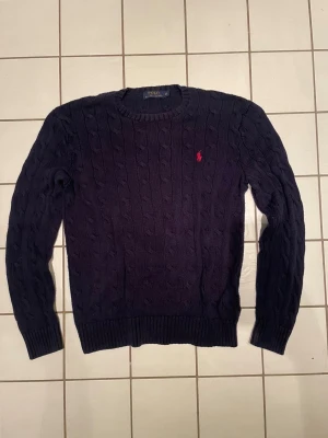 Kabelstickad tröja från Ralph Lauren - Snygg marinblå kabelstickad tröja från Polo Ralph Lauren i storlek XS. Tröjan har rund hals, långa ärmar och den klassiska röda Polo-loggan broderad på bröstet. Perfekt för dig som gillar stilrena och tidlösa plagg.