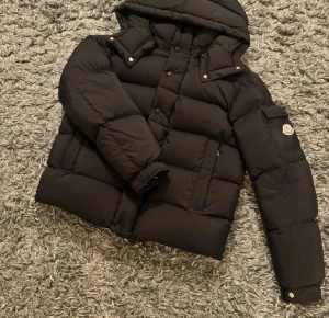 Svart Moncler pufferjacka med huva - Svart pufferjacka från Moncler med huva, stora fickor och ikonisk logga på ärmen. Jackan har både dragkedja och knappar framtill samt ribbade muddar vid ärmslut. Perfekt för kalla dagar och har en clean, modern look.