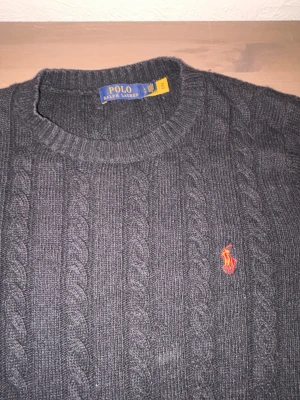 Svart stickad tröja Polo Ralph Lauren - Mörkgrå kabelstickad tröja från Polo Ralph Lauren i storlek L. Tröjan har rund hals, lång ärm och klassisk röd broderad logga på bröstet. Materialet är mjukt och stickat, perfekt för kyliga dagar. Snygg ribbad kant vid hals, ärmslut och nederkant.