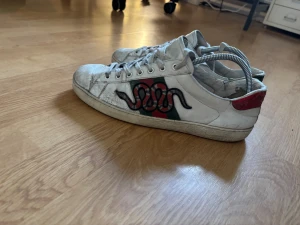 Gucci Ace sneakers med orm brodyr - Gucci Ace sneakers i vitt skinn med ikoniska gröna och röda ränder på sidan och broderad orm. Baksidan har metallicrött och grönt parti. Klassisk låg modell med snörning och rund tå. Snygga detaljer och lyxig känsla. Väldigt dåligt skick på skorna där av kan priset diskuteras. Perfekt för dig som vill göra dem i bättre skick och sälja dyrare 