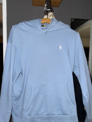 Ljusblå hoodie från Polo Ralph Lauren - Snygg ljusblå hoodie från Polo Ralph Lauren med klassisk vit logga broderad på bröstet. Tröjan har huva med snörning, stor magficka och långa ärmar. Perfekt för chill dagar och enkel att matcha med jeans eller joggers. Som ny