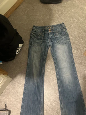 Blå jeans från Clockhouse med raka ben - Snygga blå jeans från Clockhouse med raka ben och klassisk femficksdesign. Jeansen har dubbla knappar i midjan, slitningar och ljusare tvättade partier för en cool look. Materialet är jeans och passformen är normal. Perfekt för dig som gillar avslappnad stil. Midja tvärsöver är 35 cm och innerbenslängden är 77 cm. 