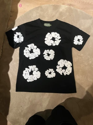 Svart  Denim Tears - Svart t-shirt från Denim Tears i storlek S med stora vita blommönster tryckta över hela plagget, både fram och bak. Rund hals och klassisk passform. Materialet är mjuk bomull och designen är riktigt unik och sticker ut. Storlek S