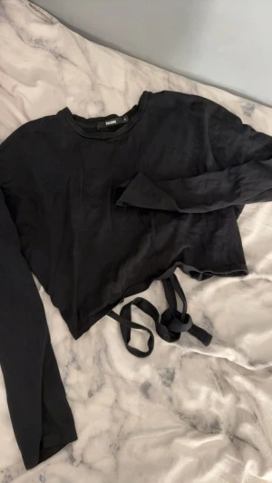 Svart croppad topp med knyt från Bik Bok - Svart långärmad croppad topp från Bik Bok med rund hals och snygga knytband i midjan. Tunn och mjuk i bomullsmaterial, perfekt för att styla med höga jeans eller kjol. Enkel design med cool edge tack vare knytet framtill.