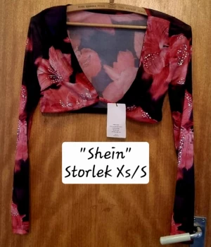 Blommig croppad bolerotopp från Shein - Säljer en snygg croppad bolerotopp från Shein med blommigt mönster i rött och rosa på svart bakgrund. Toppen har lång ärm och djup v-ringning, perfekt för att lyfta din outfit med lite färg och mönster.