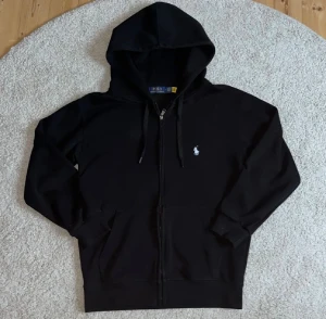 Svart hoodie från Polo Ralph Lauren - Svart hoodie från Polo Ralph Lauren med klassisk broderad logga på bröstet. Tröjan har dragkedja framtill, huva med snörning och två fickor. Tillverkad i mjuk bomull, perfekt för chill dagar eller när du vill ha en clean look. Heltintill helt ny och använd 1 gång skickas samma dag som köper har gjorts bjud kan tas emot kvitto medföljer 