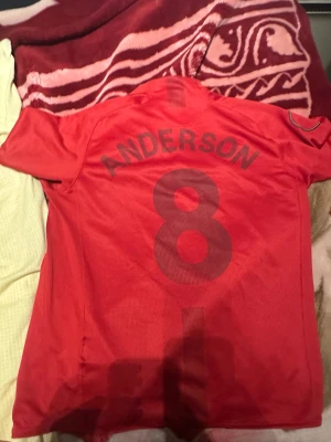 Manchester United Anderson 8 Nike tröja - Klassisk röd Manchester United matchtröja med Anderson och nummer 8 på ryggen. Broderat klubbmärke, Nike swoosh och AIG sponsorlogga på bröstet. Tillverkad i lätt och ventilerande material, perfekt för fotboll eller samling.