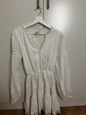 Vit spetsklänning från Nelly - Superfin vit kortklänning från Nelly med broderade spetsdetaljer och v-ringning. Klänningen har långa ärmar, markerad midja och volangkant nertill. Perfekt för sommarens alla tillfällen och ger en romantisk vibe.