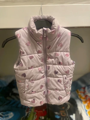 Dunväst med hjärtan från H&M - Söt ljusrosa dunväst från H&M med glansiga rosa hjärtan över hela västen. Västen har hög krage, två fickor och dragkedja framtill. Perfekt för att addera lite extra färg och värme till din outfit. Barn till tjejer, storlek 110/116