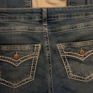 Blå jeans med kontrastsömmar - Snygga blå jeans med tydliga vita kontrastsömmar och dekorativa bakfickor med knappar. Klassisk femficksmodell och normal passform. Jeansen har medelhög midja och är tillverkade i jeansmaterial med lätt tvättad look.