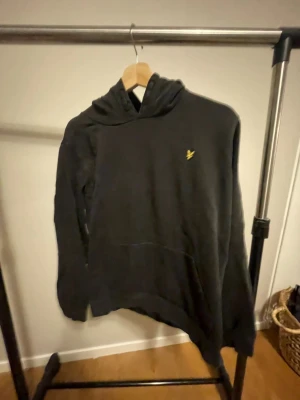 Svart hoodie från Lyle & Scott - Svart hoodie från Lyle & Scott med klassisk gul broderad logga på bröstet. Tröjan har huva, känguruficka och långa ärmar. Perfekt för dig som gillar stilrena och enkla plagg med en touch av street. Mjuk och skön bomullskänsla.