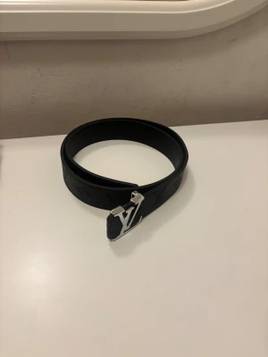 Louis Vuitton svart skärp i skinn - Snyggt svart Louis Vuitton-bälte i skinn med klassiskt LV-monogram präglat längs hela bandet. Bältet har en bred design och en silverfärgad LV-logga som spänne. Perfekt accessoar för att lyfta din outfit med en lyxig touch.