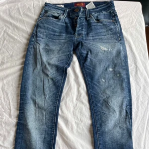 Slim Glenn jeans Jack & Jones - Snygga blå jeans från Jack & Jones, modell Slim Glenn. De har en slim passform, klassisk femficksdesign och slitna detaljer framtill för en cool look. Jeansen är tillverkade i jeansmaterial med knappgylf och kontrastsömmar. Perfekt för dig som gillar en modern stil. Storlek W27L30 och i fint skick.