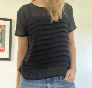 Svart blus med knappar bak - Säljer en svart blus med korta ärmar och rund hals. Framsidan har snygga horisontella volangdetaljer och baksidan är försedd med knappar. Blusen är lätt transparent och har en loose passform, perfekt att styla med jeans eller kjol.