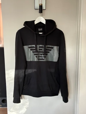  EA7 hoodie med logga - Svart hoodie från EA7 Emporio Armani med stor logga och ikonisk örn på bröstet i grått. Tröjan har huva med snörning, ribbade muddar och ficka framtill. Snyggt tryck även på ärmen. Perfekt för dig som gillar streetwear och vill ha en stilren look.