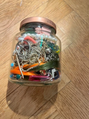 Burk med blandade smycken 546g - En mystery glasburk fylld med blandade smycken, inklusive halsband, armband, ringar och örhängen. Färgerna varierar från silver, guld, orange, grön, rosa, blå och pärlvitt. Materialen är blandade, med inslag av metall, plast och pärlor. Perfekt för dig som gillar att mixa och matcha accessoarer.